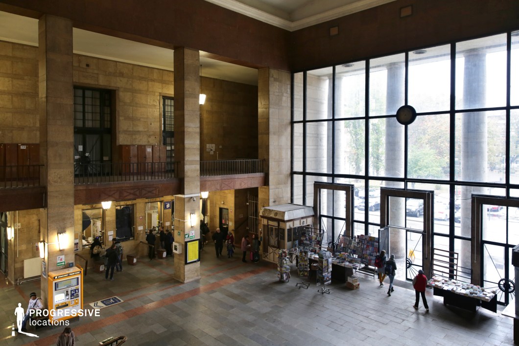 This spacious railway hall features towering stone columns and an expansive glass facade, allowing natural light to flood the interior while showcasing the imposing classical columns outside. The architecture combines grand proportions with functional elements, including a second-floor balcony and a ticketing area below. Warm stone walls and dark metal railings give the space a formal yet slightly aged appearance, while the checkered stone floor enhances the geometric aesthetic. The presence of kiosks and stalls adds a sense of daily activity. This setting is ideal for period dramas, historical narratives, or suspenseful thrillers requiring a transit hub with a timeless, bureaucratic, or nostalgic atmosphere.