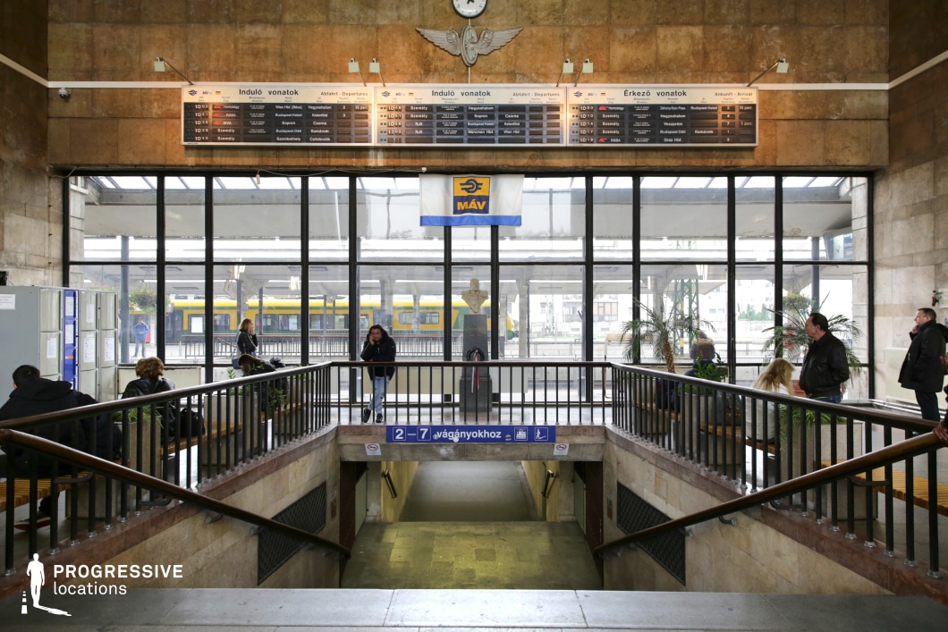 This train station interior features a grand staircase leading to the underground passageways, framed by dark metal railings and marble-textured walls. The architecture blends mid-century modern and functionalist styles, with warm-toned stone cladding on the walls and large industrial-style windows providing a view of the platform and a passing train. Above the entrance, classic split-flap timetable boards display arrivals and departures, enhancing the nostalgic atmosphere. The space is illuminated by both natural light and retro wall-mounted lamps, reinforcing the old-world charm. This setting is ideal for filming scenes requiring a transit hub with a timeless, slightly worn aesthetic. It works particularly well for drama, period films, thrillers, or stories emphasizing travel, farewells, and personal transitions.