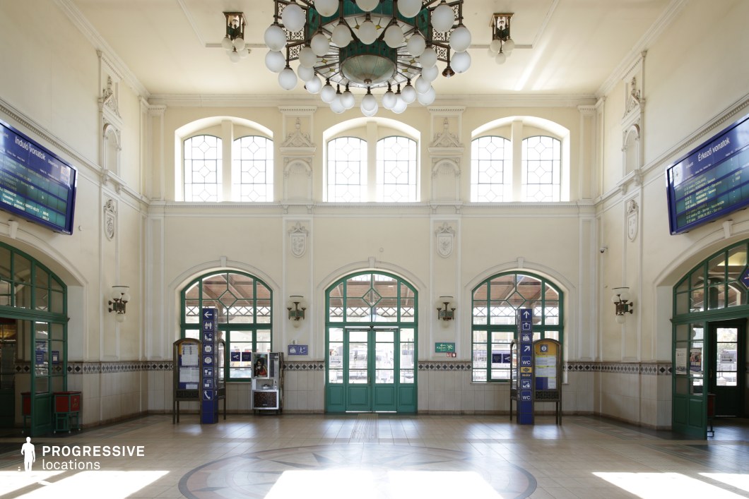 This image shows the interior of a historic train station, with a grand architectural design. The space is bathed in natural light from the large, arched windows that span across the top of the room. A majestic chandelier hangs from the ceiling, made up of numerous globe-shaped light fixtures, adding an ornate touch. The walls feature detailed moldings and decorative elements, while the floor is covered with elegant, geometric tiles. On either side of the room, green-framed doors open to the outside, and signage on posts directs towards amenities like restrooms and train schedules. The combination of classical architectural features and modern functional elements gives the space a timeless charm.