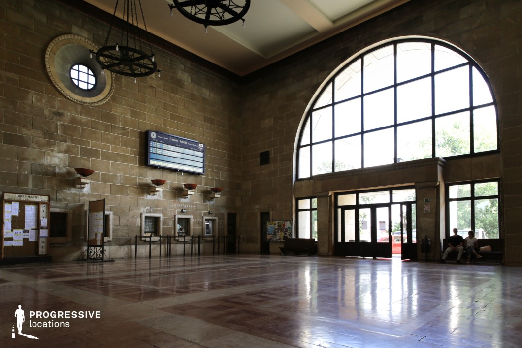This spacious train station waiting hall features high ceilings, large arched windows, and a polished marble floor that reflects the incoming natural light. The architecture is a blend of early 20th-century industrial and classical elements, with textured brick walls, heavy iron chandeliers, and vintage ticket counters embedded in the stone façade. A circular window and metal-framed glass doors add to the symmetrical design, while notice boards and a retro departure board reinforce the functional purpose of the space. This location is ideal for filming historical dramas, period pieces, and contemplative or departure scenes, offering a setting that evokes nostalgia, grandeur, and quiet anticipation.