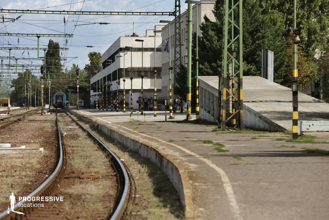 This train station features a distinctly Soviet-era architectural design, with a functional, rectangular main building that includes overhanging upper floors and tinted windows. The platform is lined with tall, green metal poles supporting a complex network of overhead electric lines, creating a structured and industrial visual. A gently curving railway track leads towards the horizon, adding depth and perspective. The worn concrete, weathered surfaces, and yellow-black safety markings enhance the station’s utilitarian, slightly aged aesthetic. This location is ideal for filming Cold War-era period pieces, industrial dramas, or suspenseful scenes involving travel, espionage, or departures.