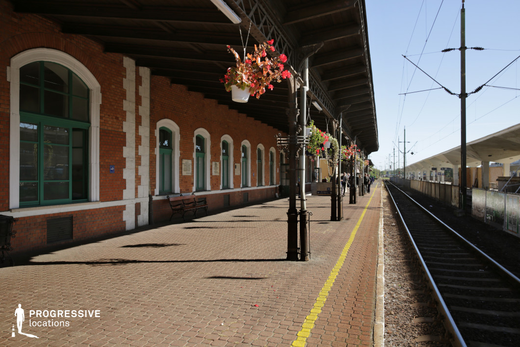 This scenic train platform features a charming mix of brick walls and intricate ironwork. The sturdy, red-brick façade is complemented by tall, arched windows with green frames, offering a glimpse of the station's vintage design. The platform is enhanced with hanging flower baskets, bringing vibrant colors and a touch of nature to the industrial surroundings. The benches along the platform, along with the textured paving, contribute to the welcoming ambiance. The clear view of the tracks and overhead electrical lines creates a functional yet visually appealing setting, ideal for capturing the essence of a timeless transportation hub.
