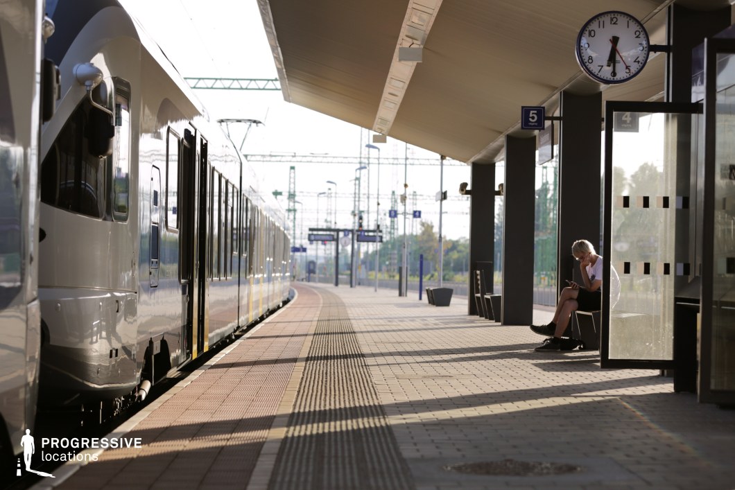 This contemporary train platform features a sleek, streamlined design with a polished overhead canopy, dark metal support columns, and a smooth tiled surface. A modern train with reflective surfaces extends into the distance, emphasizing depth and movement. The station is equipped with clear signage, a prominent round clock, and well-organized waiting areas, enhancing its efficient, minimalist aesthetic. The interplay of light and shadow creates a dynamic composition, adding visual interest. This location is ideal for filming travel sequences, introspective moments, or scenes set in a transitional space, making it suitable for dramas, thrillers, and futuristic or urban-themed narratives.