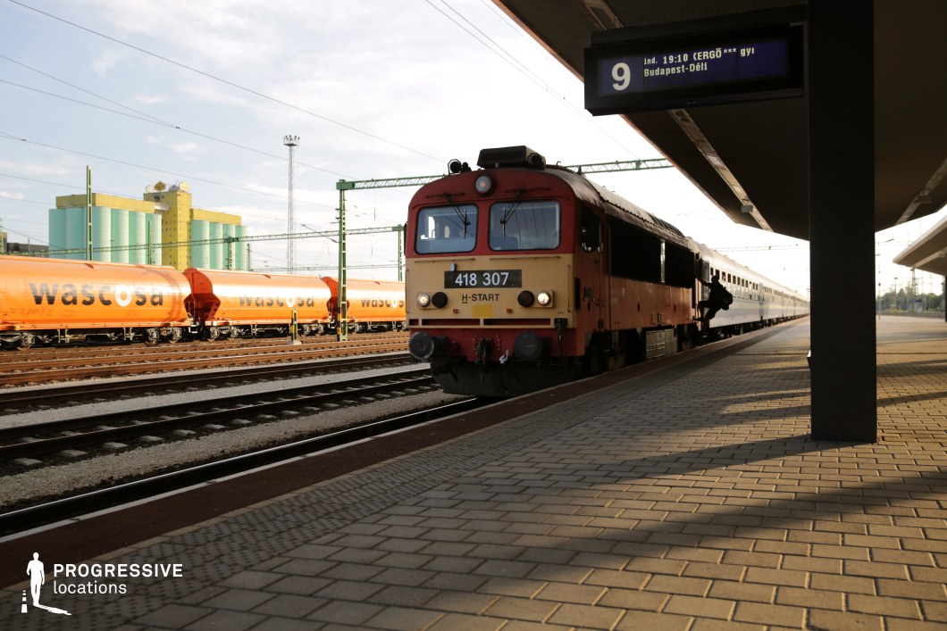 This train platform features a vintage red-and-yellow diesel locomotive pulling a set of passenger cars, framed by an industrial railway yard. Overhead electronic signage provides departure details, reinforcing the sense of movement and transit. In the background, bright orange freight wagons and large pastel-colored grain silos create a visually striking industrial contrast. The platform's covered walkway casts long, angular shadows, adding depth and visual interest to the scene. This location is ideal for filming travel-based narratives, industrial backdrops, or period scenes, making it well-suited for dramas, thrillers, and historical films set in transportation hubs.
