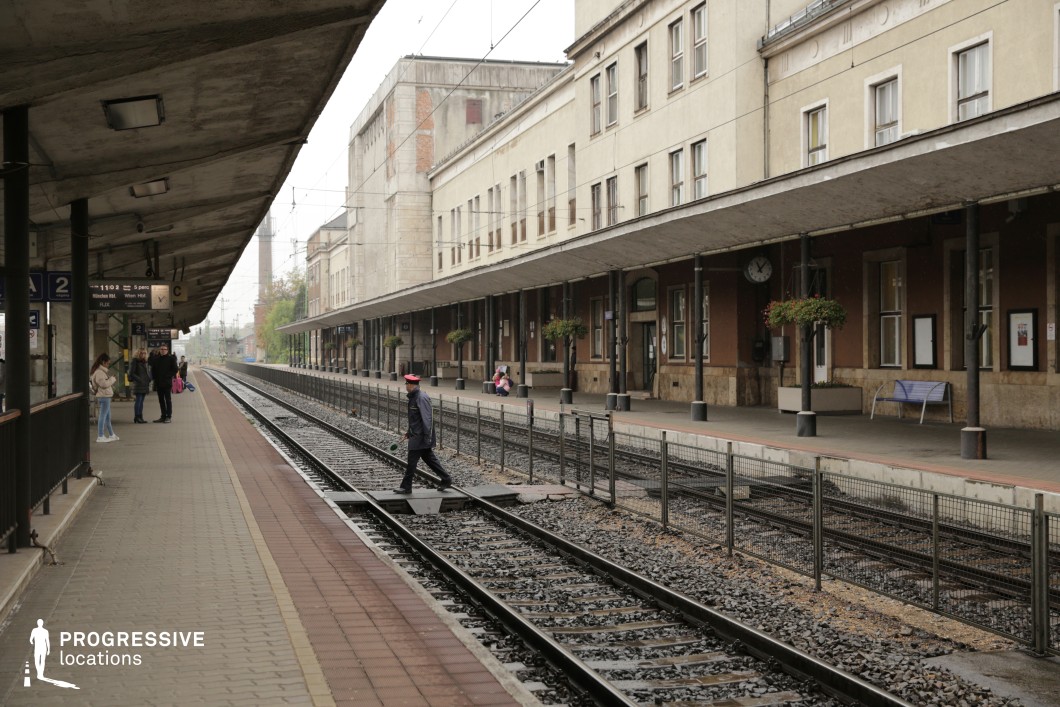 This mid-20th-century railway station features long, covered platforms extending parallel to multiple train tracks. The architecture is functional and slightly worn, with a mix of concrete, metal, and brickwork, giving it a utilitarian, slightly aged character. Overhanging concrete canopies supported by black metal columns provide shelter, while the station building has a linear, repetitive facade with numerous windows and subtle decorative elements. The tracks, flanked by metal railings, stretch into the distance, creating a strong sense of depth and perspective. The slightly weathered appearance and muted color palette make this an excellent filming location for period dramas, crime thrillers, or stories with a sense of transience, departure, or nostalgia.