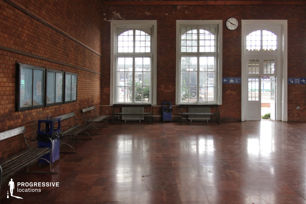 This historic waiting area features exposed red brick walls and large arched windows framed in white, allowing natural light to filter in. A polished tile floor reflects the light, enhancing the room’s depth and texture. Metal benches with a simple, utilitarian design line the walls, accompanied by radiators beneath the windows. The high ceiling and period details, such as the wall-mounted clock and signage, contribute to the station's timeless appeal. This setting is ideal for classic travel scenes, period dramas, or projects evoking a sense of nostalgia and transit history.