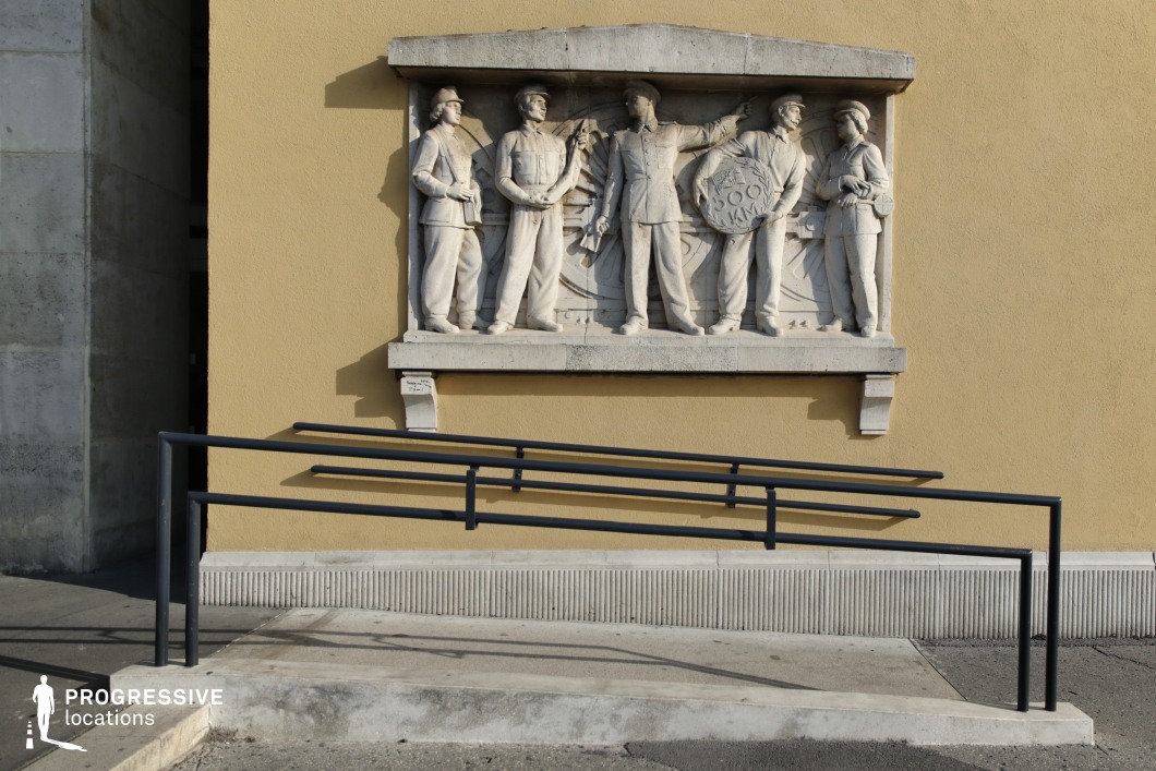 This architectural relief, set against a yellow-toned plaster wall, depicts a sculpted group in a socialist-realist style, symbolizing industrial labor and progress. The figures are carved from light-colored stone, standing in front of a mechanical backdrop, adding depth to the composition. Below, a modern black metal handrail and accessibility ramp contrast with the historical relief, creating an interesting juxtaposition between past and present. The setting offers a visually compelling location for filming, ideal for historical dramas, political thrillers, or documentary-style narratives exploring industrial heritage, social movements, or architectural evolution.