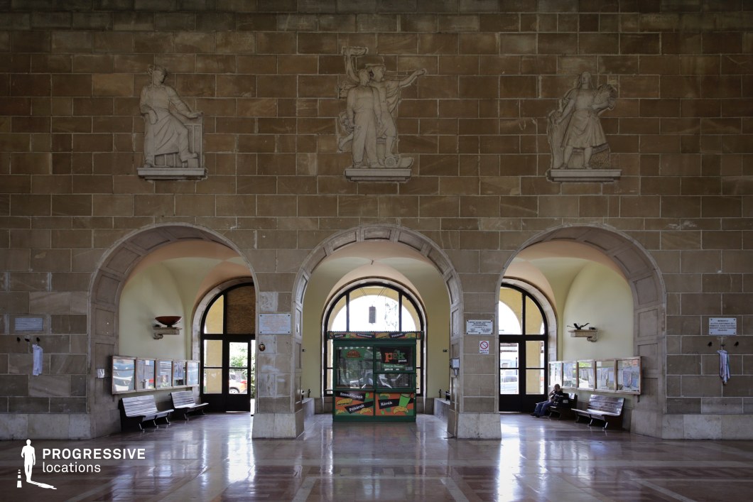 This train station waiting hall features imposing arched passageways framed by textured stone walls and adorned with classical relief sculptures depicting allegorical figures. The polished marble floor reflects the ambient light, enhancing the sense of grandeur and permanence. The arched doorways lead to separate seating areas with wooden benches, while large, curved windows allow natural light to filter into the space. The symmetrical design and historical details evoke a sense of old-world formality and institutional authority. This location is ideal for filming period dramas, historical documentaries, or scenes requiring a setting with a grand, slightly austere atmosphere, making it suitable for themes of bureaucracy, transition, or nostalgia.