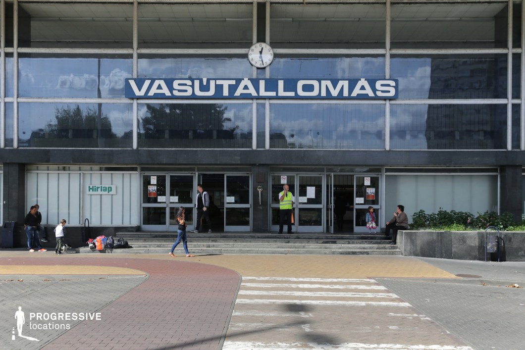 This train station entrance features a bold mid-century modernist design, characterized by large glass windows, a prominent station name in retro block lettering, and a central round clock mounted above the entrance. The dark stone-clad exterior contrasts with the reflective glass panels, which capture the surrounding urban environment. A set of symmetrical doors and steps lead to the main hall, while signage and architectural details reinforce the functional, utilitarian aesthetic. This location is ideal for filming travel-related sequences, urban dramas, or period pieces set in the mid-to-late 20th century, offering a sense of nostalgia, movement, and public transit atmosphere