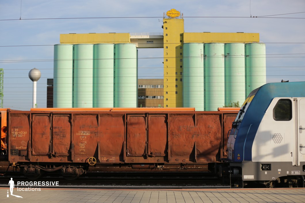 This train yard setting juxtaposes industrial elements with vibrant architectural features. A rusted freight wagon and a sleek modern locomotive dominate the foreground, reinforcing the contrast between aging infrastructure and contemporary rail transport. In the background, pastel green and bright yellow silos tower over the scene, part of a large industrial grain storage facility. The geometric repetition of the cylindrical silos and the vertical structure adds depth and symmetry, while the bold colors make the space visually distinctive. This location is ideal for filming scenes that emphasize industrial landscapes, economic transitions, or transport hubs, making it well-suited for documentaries, thrillers, and dystopian or post-industrial narratives.