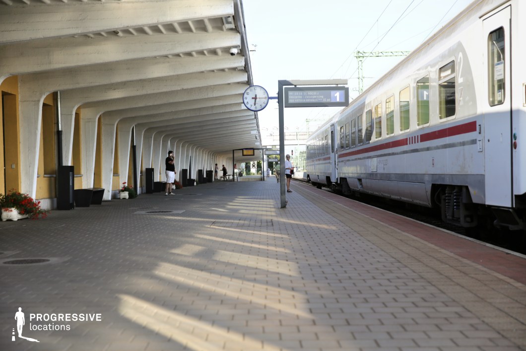 This train platform features a striking mid-century modern canopy with repetitive, curved concrete beams that create a rhythmic visual pattern. The shelter, supported by angular white columns, contrasts with the yellow station building, adding warmth to the scene. The paved platform extends towards the horizon, where an arriving train with sleek white carriages and red detailing reinforces the sense of movement and transit. A large, round station clock and electronic signage contribute to the functional, slightly retro-futuristic aesthetic. This location is ideal for filming scenes centered around departures, arrivals, and travel-related tension, making it well-suited for dramas, thrillers, and nostalgic period pieces.