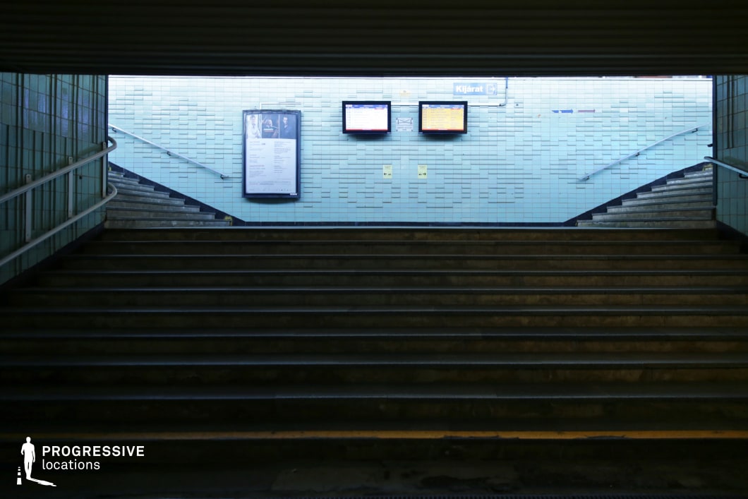 This underground passage features a dark, aged staircase leading upward to a brightly lit exit, framed by cool-toned blue tiles that create a stark contrast between the confined lower space and the openness above. Metal railings line both staircases, reinforcing the structured, functional design. Overhead electronic display boards provide transit information, adding a practical yet slightly outdated aesthetic. The combination of artificial lighting and worn surfaces contributes to a moody, slightly eerie atmosphere. This location is ideal for filming tense, suspenseful scenes, making it well-suited for thrillers, urban dramas, or noir-style cinematography emphasizing isolation, secrecy, or transition.