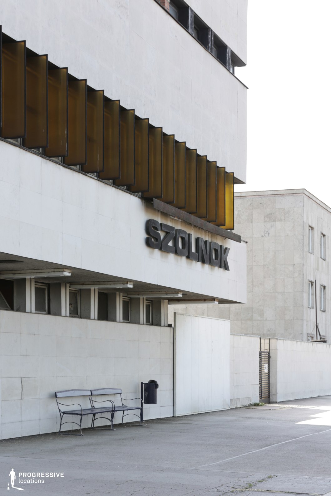 This imposing mid-century modernist train station facade is characterized by stark concrete walls, bold block lettering, and a distinctive row of amber-tinted vertical window shades. The minimalist, geometric aesthetic is reinforced by the building's clean lines and rigid, functional design. The surrounding environment, with a simple bench and metal trash bin, emphasizes the utilitarian atmosphere of the space. The slightly weathered appearance of the materials adds to its historical, almost dystopian feel. This location is ideal for filming Cold War-era narratives, noir thrillers, or gritty urban dramas that require a setting with strong architectural presence and an air of isolation or bureaucracy.