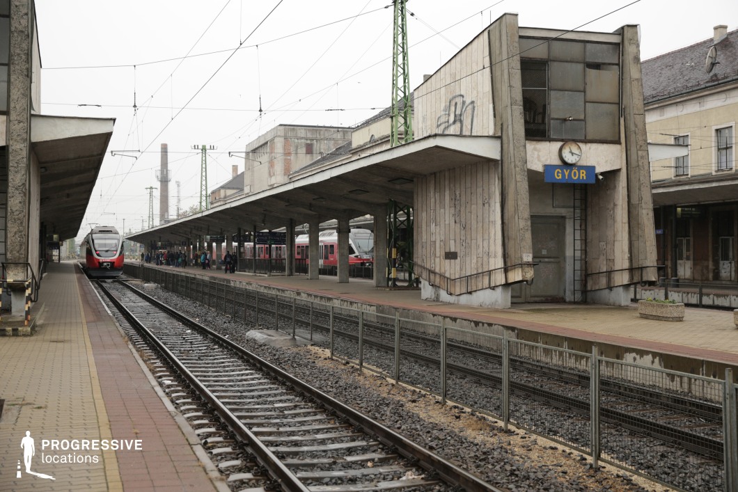This railway station blends utilitarian and brutalist architecture, characterized by raw concrete structures, angular overhangs, and a starkly functional design. The weathered central platform building, with its slanted concrete walls and metal-framed windows, stands as a focal point, while the extended covered walkways emphasize depth and perspective. Overhead electric wires and industrial elements, such as green steel pylons and distant chimneys, reinforce the station’s gritty, mid-century atmosphere. The presence of modern red and white trains contrasts with the aged infrastructure, highlighting a mix of past and present. This setting is ideal for period dramas, dystopian thrillers, or Cold War-era films requiring a raw, institutional backdrop with a sense of historical tension.