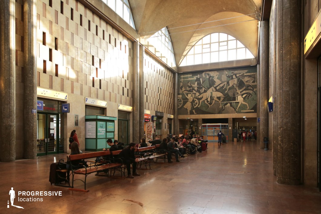This grand train station waiting hall features soaring arched ceilings, tall marble-clad columns, and an expansive mosaic mural depicting a dynamic socialist-era scene of workers and industry. The geometric wall pattern, combined with the rich textures of stone and metal, reflects mid-20th-century monumental architecture. Large arched windows allow natural light to filter in, enhancing the dramatic atmosphere. Wooden benches provide seating beneath the high ceilings, reinforcing the scale of the space. This location is ideal for filming historical and political dramas, Soviet-era period pieces, or scenes emphasizing institutional grandeur, transit, and social movements.