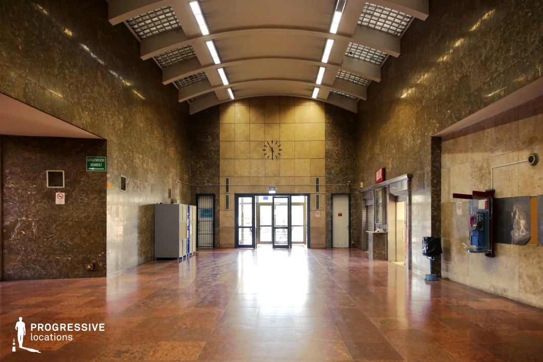 This train station waiting hall features a striking mid-century modernist design, characterized by dark marble-clad walls, a curved ceiling with integrated lighting panels, and polished reddish-brown flooring. The symmetrical layout is centered around a minimalist clock and a set of glass doors leading to the exterior, enhancing the hall’s functional yet imposing atmosphere. Utility elements, such as an old payphone, metal lockers, and shuttered service windows, reinforce the slightly austere, institutional feel of the space. This location is ideal for filming historical dramas, Cold War-era thrillers, or dystopian narratives, making use of its monumental scale, moody lighting, and rigid architectural lines.