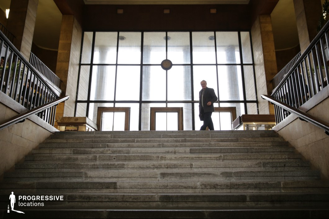 This grand interior space features a wide, imposing staircase made of polished stone, leading up to a striking glass entrance framed by tall, classical columns. The symmetrical design is accentuated by dark metal railings on both sides, creating a structured and formal aesthetic. The large glass facade allows natural light to flood the space, contrasting with the warm beige tones of the surrounding stone walls. The atmosphere is monumental and slightly austere, making this an excellent setting for political dramas, historical films, or noir thrillers that require a setting with authority, power, or institutional grandeur.