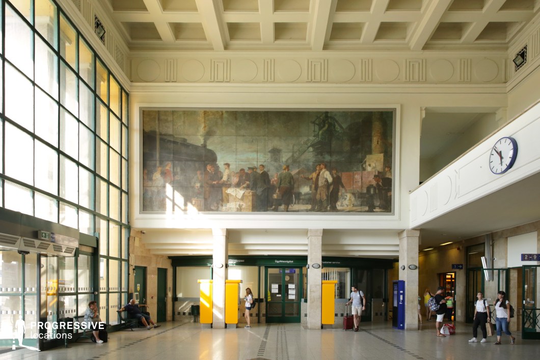 This spacious train station hall features a high ceiling with intricate geometric detailing and large glass-paneled walls, allowing abundant natural light to flood the space. The focal point is a massive mural above the main entrance, depicting an industrial-era scene in muted, earthy tones, adding historical significance to the environment. The hall combines classical and modernist elements, with polished stone flooring, sturdy columns, and functional ticketing and customer service areas. The contrast between the monumental artwork and the streamlined architectural design makes this an ideal filming location for period dramas, political thrillers, or narratives exploring history, bureaucracy, or personal departures and reunions.