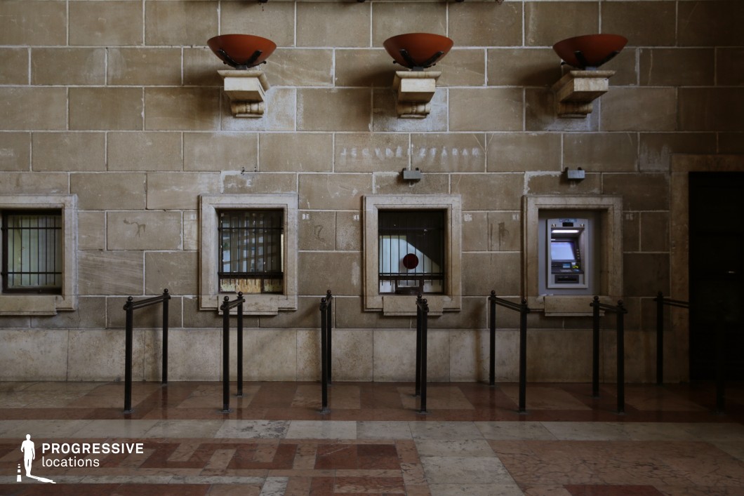 This vintage ticket counter is set against a textured stone wall, featuring small, barred service windows framed by classical stone borders. Above, decorative sconces with rust-colored bowls add an old-world aesthetic, while the worn marble flooring reflects years of foot traffic. The dark metal queuing barriers enhance the structured, formal design, leading toward the once-active service points. A single modern ATM contrasts with the aged surroundings, highlighting the passage of time. This location is ideal for filming historical dramas, noir thrillers, or atmospheric urban stories, emphasizing themes of nostalgia, bureaucracy, or abandonment.