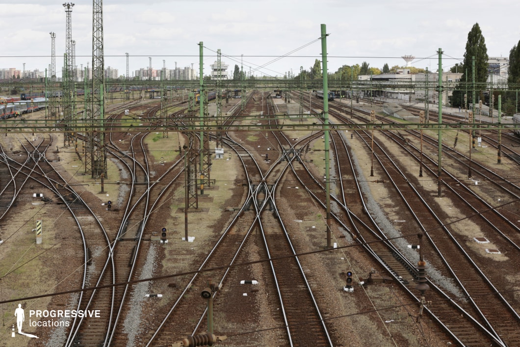 This vast rail yard features a complex web of railway tracks, crisscrossing and diverging in multiple directions. Overhead electrical lines and towering metal poles create a dense industrial landscape, with green-painted supports adding a structured rhythm to the scene. The worn brown and gray gravel between the rails, along with patches of dry grass, gives the location an aged, utilitarian feel. The distant skyline of residential and industrial buildings adds depth, reinforcing the setting as a major transit hub. This location is perfect for filming logistical operations, crime thrillers, or historical films set in the mid-to-late 20th century, emphasizing movement, tension, or clandestine activities.