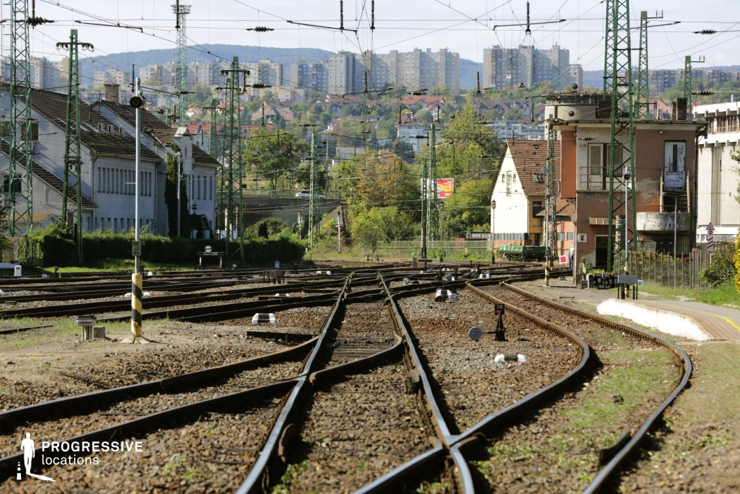 This image captures a view of a train yard with intersecting tracks leading through the scene. The railway lines wind through the landscape, flanked by a mix of industrial buildings and residential complexes in the background, offering a sharp contrast between the aging infrastructure and modern city life. Overhead power lines stretch above, adding to the industrial feel of the location. A small platform, marked with a yellow line, sits in the foreground, with a building featuring a worn façade positioned to the right. The city skyline looms in the distance, emphasizing the urban setting.