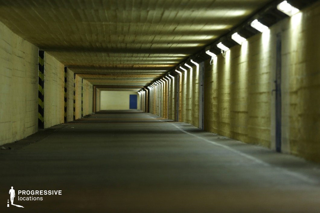This long, industrial underpass is constructed from raw concrete and illuminated by a row of harsh fluorescent lights, casting a stark contrast against the dark ceiling and floor. The repetitive beams and structural supports create a strong sense of depth and perspective, with a small blue door at the far end acting as a focal point. The worn, utilitarian aesthetic, coupled with the muted yellowish tones, gives the space an eerie, almost dystopian feel. This setting is ideal for filming suspenseful scenes, chase sequences, or psychological thrillers where a confined, oppressive atmosphere is required.