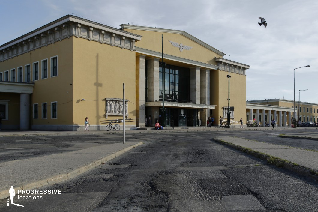 This imposing neoclassical railway station features a symmetrical facade with a bold yellow exterior contrasted by white decorative elements. Tall, evenly spaced columns frame the entrance, while sculpted reliefs adorn the walls, adding historical and artistic depth. The grand entrance, emphasized by a glass doorway and an overhanging canopy, enhances the building’s monumental feel. The foreground showcases a weathered, cracked asphalt road leading up to the structure, contributing to an urban, slightly gritty atmosphere. The linear design and repetitive colonnades create a strong visual rhythm, making this location ideal for period films, political dramas, or urban thrillers where an institutional or authoritarian aesthetic is needed.