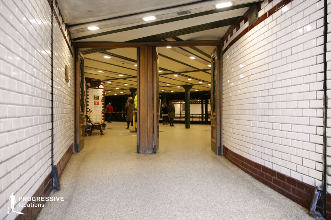 This image captures the entrance to a historic underground metro station, framed by white subway tiles with red trim, creating a clean yet vintage aesthetic. The doorway, featuring wooden double doors slightly ajar, leads into a well-lit station with dark green riveted iron columns and an exposed-beam ceiling. The polished neutral flooring reflects the overhead lights, enhancing the sense of depth. The structured symmetry and blend of old-world charm with functional urban design make this location ideal for filming period dramas, classic European city sequences, or introspective urban moments, offering a timeless transit setting.