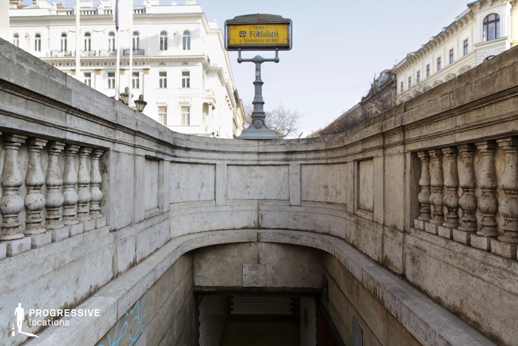 This image showcases an elegant underground metro entrance framed by a grand stone balustrade with classical detailing. The aged stone surface, with its weathered texture, adds a sense of history and character. A vintage-style yellow metro sign atop an ornate metal post provides a focal point, while the entrance itself leads down into a dimly lit tiled passageway. In the background, neoclassical buildings with decorative balconies and arched windows further enhance the European architectural charm. This location is ideal for filming period dramas, historical urban sequences, or atmospheric noir scenes, offering a refined and cinematic city backdrop.
