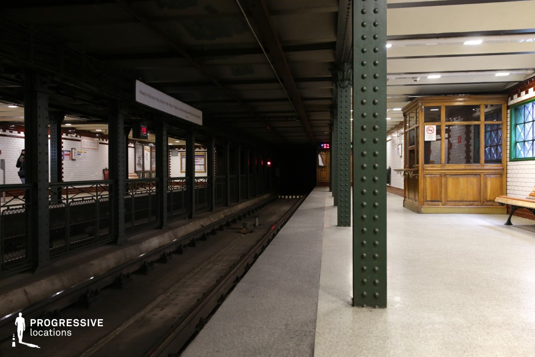This image showcases a vintage metro platform with green riveted iron columns and a dark, tunnel-like track leading into the distance. The station's walls are adorned with white ceramic tiles and brown brick trim, evoking an early 20th-century architectural style. A wooden ticket booth with glass panels and a decorative counter adds to the nostalgic charm, while a bench provides a functional waiting area. The dim lighting along the tracks contrasts with the well-lit platform, creating a visually striking scene. This location is ideal for historical film productions, noir-style cinematography, or period-set urban storytelling.