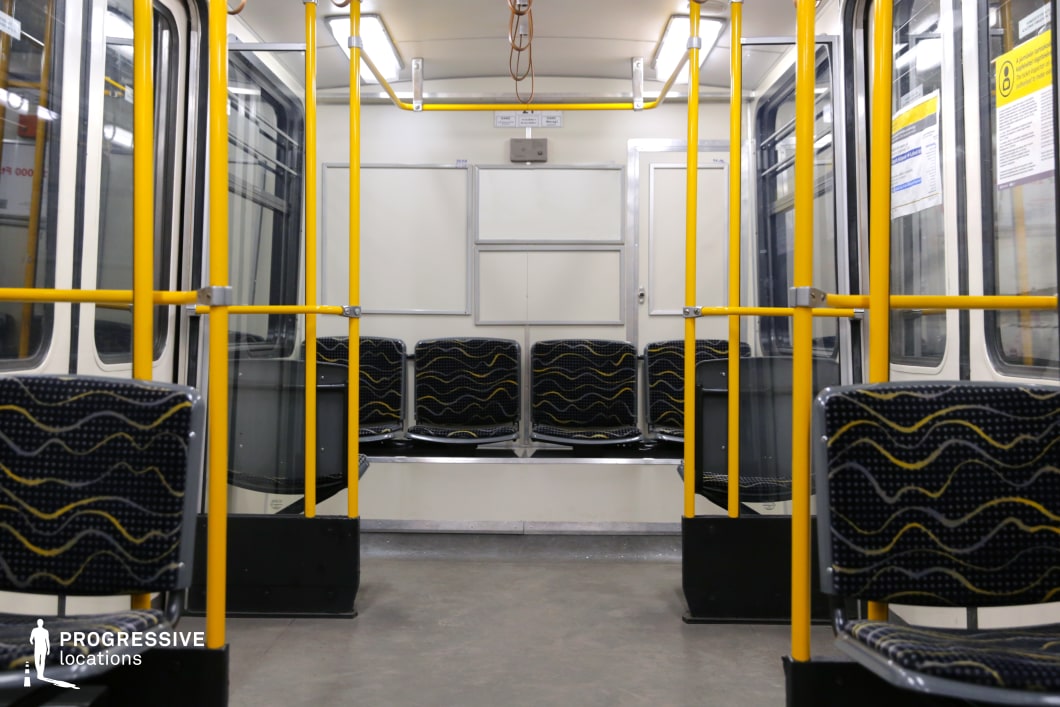 This image captures the interior of a classic metro train carriage, characterized by its retro design and functional layout. The seating arrangement consists of black patterned fabric seats with yellow wavy accents, arranged along the sides to maximize space. Bright yellow support poles and handrails contrast with the neutral white and gray tones of the walls and flooring. Overhead fluorescent lighting illuminates the space evenly, creating a clean and practical atmosphere. The windows and doors are framed with metallic elements, adding to the industrial aesthetic. This setting is ideal for film productions requiring a historical metro interior or a minimalist urban transit scene.