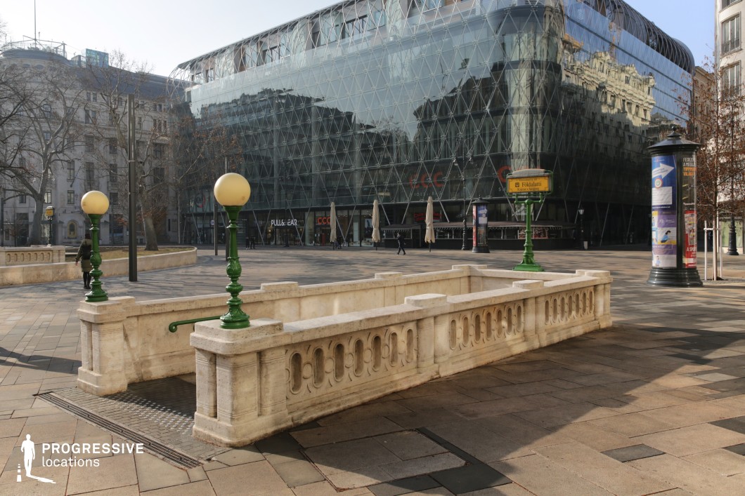 This image features a historic metro entrance with ornate stone balustrades and vintage-style green lampposts, seamlessly integrated into a contemporary urban square. The stonework exhibits classical architectural elements, contrasting with the sleek, glass-fronted modern building in the background. The entrance is marked by a yellow metro sign, adding a nostalgic touch to the scene. The surrounding paved plaza is spacious and open, offering an excellent setting for urban storytelling, fashion photography, or cinematic sequences that juxtapose history with modernity.