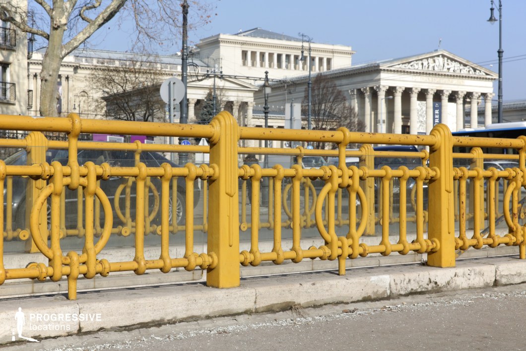 This image highlights a historic urban detail with intricate yellow wrought-iron railings, featuring elegant circular and linear patterns. The railings frame a grand neoclassical backdrop, where imposing white stone buildings with Corinthian columns and sculpted pediments dominate the skyline. The composition juxtaposes the aged, slightly weathered texture of the railings against the refined architectural grandeur beyond. This setting is ideal for film productions, historical documentaries, or urban aesthetics projects emphasizing European heritage and timeless design elements.