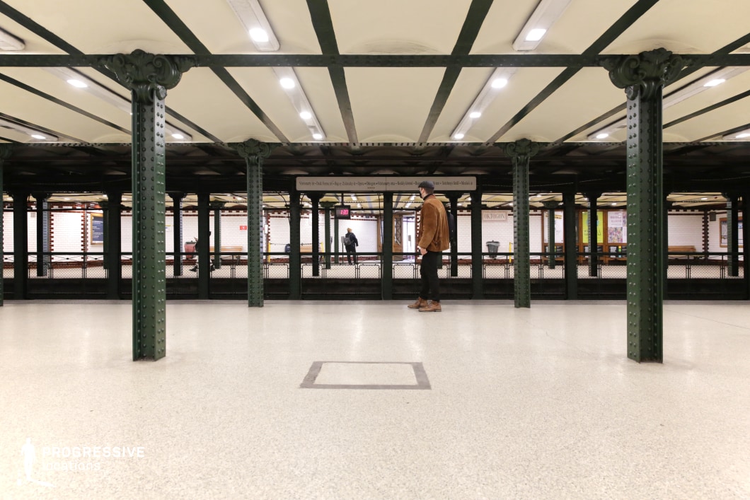 This image captures a historic underground metro station featuring a symmetrical arrangement of dark green riveted iron columns supporting a cream-colored ceiling with exposed beams. The flooring is smooth and neutral-toned, contrasting with the white tile walls adorned with red decorative trim. The open perspective emphasizes depth, with repeating columns leading toward the far end of the station. The wrought-iron railing and vintage signage reinforce the station’s early 20th-century European aesthetic. This location is ideal for filming suspenseful, noir-inspired sequences, historical narratives, or urban dramas, offering a timeless transit hub ambiance with strong visual structure.