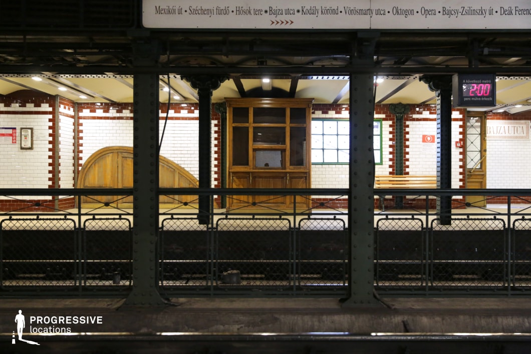 This image captures a vintage underground metro platform, featuring a classic design with white subway tiles accented by red trim. The station is framed by dark green riveted iron columns and metal railings, reinforcing its industrial-era aesthetic. A wooden ticket booth and an arched wooden door add a nostalgic charm, while a station sign and a digital countdown timer create a contrast between historical and modern elements. The composition, with its symmetrical lines and layered depth, makes this an ideal filming location for period dramas, suspenseful urban sequences, or noir-style storytelling, offering a timeless and atmospheric transit setting.