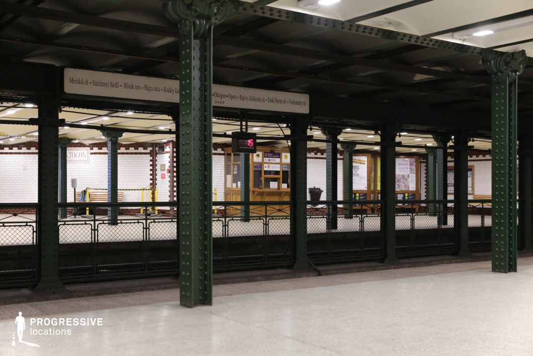 This image captures a historic underground metro station with an intricate arrangement of dark green riveted iron columns supporting an exposed-beam ceiling. The white-tiled walls, accented with red trim, contrast with the industrial metal framework. The station’s name and directional signage are displayed in a vintage-style font, reinforcing the period atmosphere. Wooden paneling and benches add warmth to the structured aesthetic, while metal railings frame the platform edge. The depth and symmetry of the scene create a visually engaging composition. This location is ideal for filming historical urban sequences, suspenseful underground scenes, or classic European transit settings, making it well-suited for noir, drama, or mystery genres.