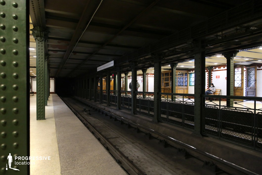This image captures a historic underground metro station, featuring dark green riveted iron columns that frame the platform and tracks. The tracks lead into a shadowy tunnel, creating a strong contrast with the well-lit opposite platform, which is adorned with white subway tiles, red trim, and wooden paneling. The metal railings and classic architectural details reinforce the vintage aesthetic of the station. The depth of the scene, combined with the interplay of light and darkness, makes this an ideal location for filming noir-style thrillers, suspenseful urban sequences, or period dramas requiring a classic European metro ambiance.