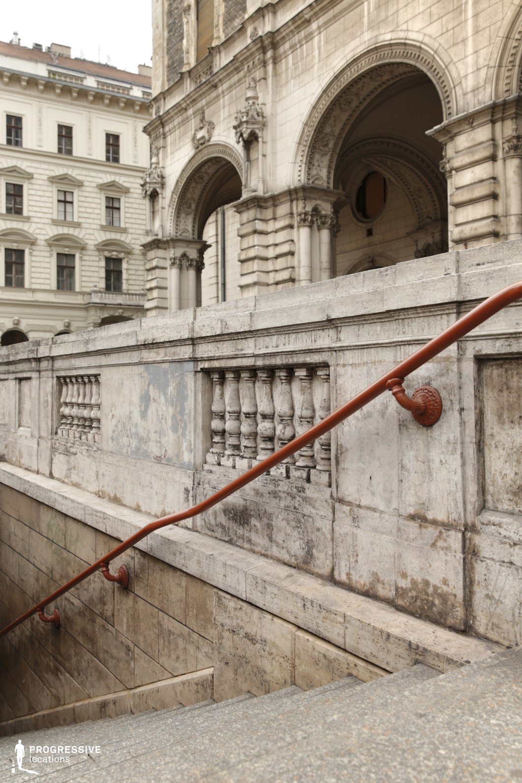 This image features an elegant stone stairway leading underground, framed by a historic balustrade with sculpted details. The aged stone surface, with its patina and subtle wear, enhances the classical character of the scene. A curved metal handrail, attached with decorative iron brackets, guides the descent. In the background, a grand neoclassical building with elaborately carved archways, intricate cornices, and sculptural elements adds depth and grandeur to the composition. This location is ideal for filming historical dramas, period pieces, or noir-style urban sequences, offering a timeless European setting with refined architectural richness.