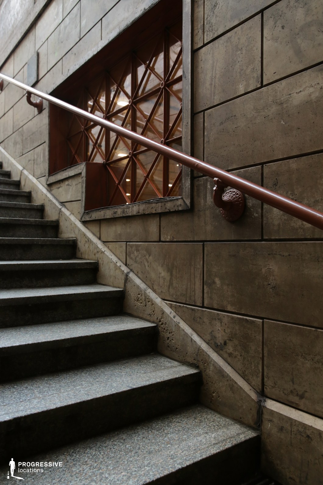 This image features a grand stone staircase leading upwards, framed by weathered stone walls with a rich patina. The polished metal handrail, mounted on intricately crafted brackets, follows the incline, adding a sense of elegance to the space. A striking window with geometric lattice detailing allows light to filter through, enhancing the old-world charm of the location. The textured stone, metal accents, and timeless architectural elements make this an ideal setting for historical dramas, suspense thrillers, or introspective character moments in film, offering a visually compelling and atmospheric backdrop.