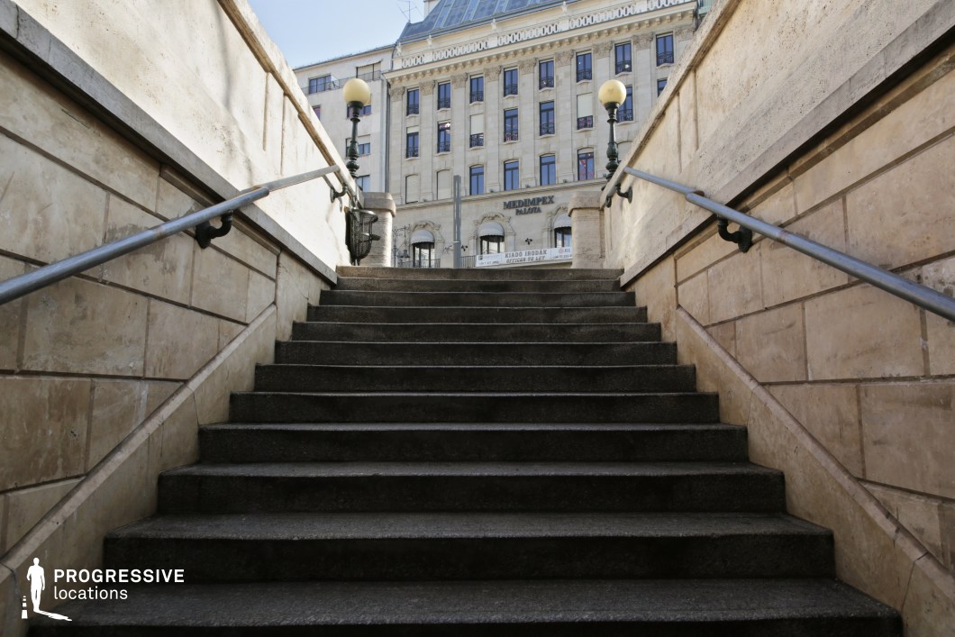 This image showcases a grand stone stairway leading up to an open plaza, framed by textured beige stone walls. Metal handrails with ornate iron brackets run along both sides, adding to the historic character of the setting. Decorative street lamps with spherical globes stand at the top of the stairs, enhancing the refined atmosphere. In the background, a stately neoclassical building with large windows and an intricate facade contributes to the European architectural elegance. The upward perspective emphasizes depth and grandeur, making this location ideal for filming dramatic ascents, historical urban scenes, or introspective moments in period dramas or noir-style storytelling.