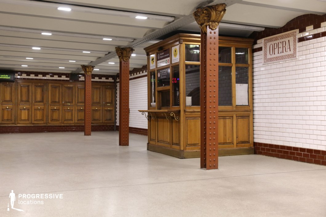 This image showcases a vintage underground metro station with a rich historical character. The space is defined by white subway-tiled walls with red trim, contrasted by warm wooden paneling and cabinetry. A classic wooden ticket booth with glass windows and ornate detailing sits centrally, evoking an early 20th-century European transit aesthetic. The riveted iron columns, painted in a deep red hue with decorative golden capitals, support an exposed-beam ceiling, reinforcing the station's period charm. The polished flooring and structured symmetry make this location ideal for filming period dramas, refined urban sequences, or atmospheric noir-style storytelling.