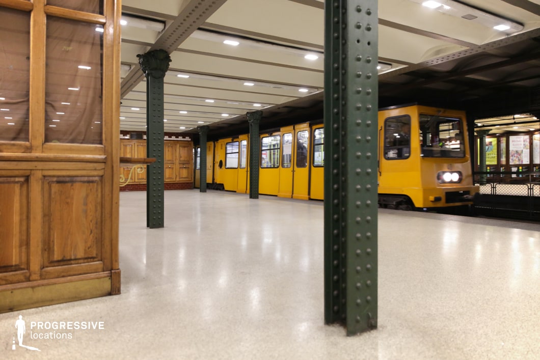 This image captures a historic underground metro station featuring a yellow train approaching the platform. The setting is characterized by riveted green iron columns supporting an exposed-beam ceiling, adding to the industrial-era charm. The platform is lined with smooth, neutral-toned flooring, while wooden ticket booths and decorative red-and-white tile accents contribute to a classic European transit aesthetic. The image’s depth and perspective emphasize the station’s structured elegance. This location is ideal for filming period dramas, suspenseful metro sequences, or urban noir scenes, offering a timeless and atmospheric underground transit setting.