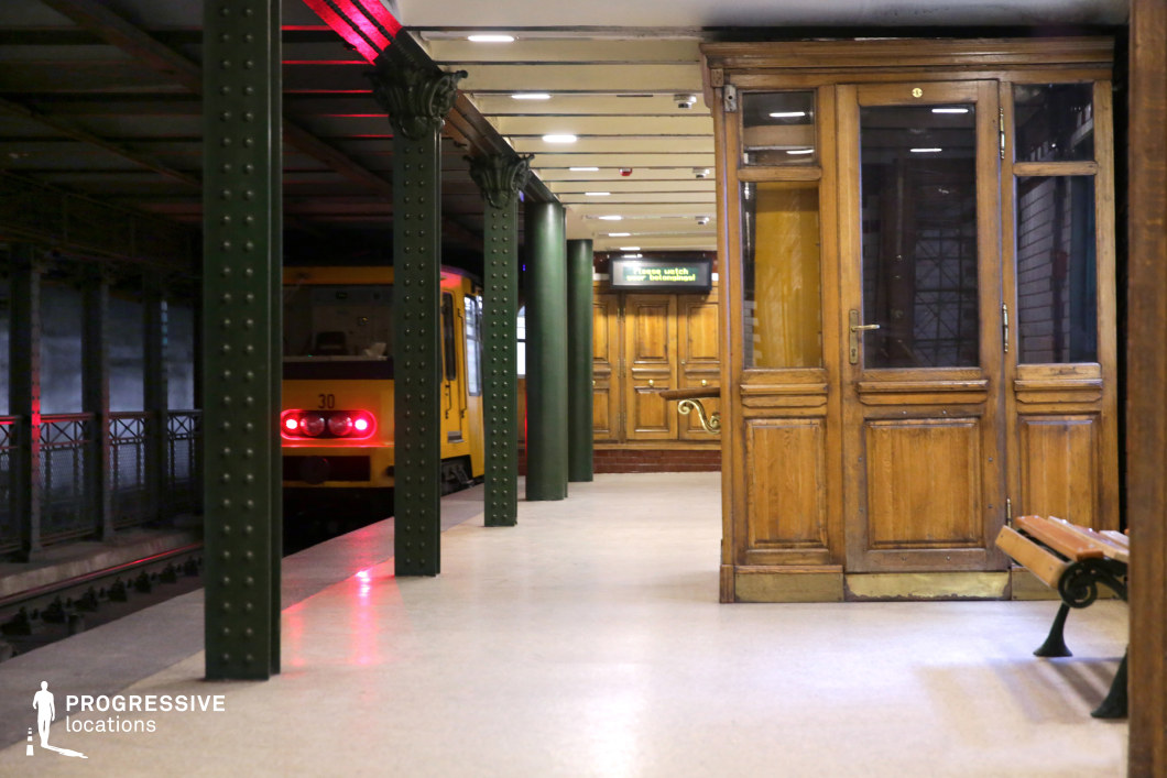 This image captures a historic underground metro platform featuring a vintage yellow train departing in the background. The setting is characterized by dark green riveted iron columns, an exposed-beam ceiling, and polished neutral flooring. A wooden ticket booth with glass panels and decorative moldings stands prominently, enhancing the station’s classic European aesthetic. Additional wooden paneling and wrought-iron railings contribute to the station’s nostalgic atmosphere. The contrast between the structured metal elements and the warm wooden details makes this an ideal location for filming period dramas, noir thrillers, or introspective urban sequences, offering a timeless transit setting.