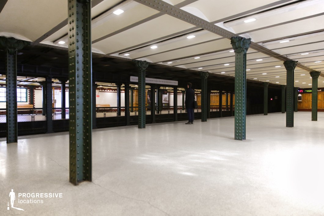 This image showcases a vintage underground metro station with a structured and symmetrical design. The space is defined by dark green riveted iron columns supporting an exposed-beam ceiling, contributing to its industrial-era charm. The platform is lined with polished neutral flooring, reflecting the overhead lighting. Across the tracks, the opposing platform features white subway tiles with red trim, wooden paneling, and classic bench seating, reinforcing the station’s historical aesthetic. The depth and repetition of architectural elements create a cinematic composition, making this location ideal for period dramas, noir thrillers, or atmospheric urban storytelling.