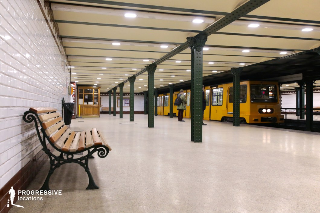This image captures a historic underground metro platform with a distinct vintage aesthetic. The platform is lined with dark green riveted iron columns supporting an exposed-beam ceiling, while the walls are clad in white subway tiles with red trim. A wooden bench with ornate wrought-iron armrests sits against the tiled wall, enhancing the station's old-world charm. A yellow metro train with a boxy design is stationed along the platform, contributing to the classic European transit feel. The symmetry of the scene, with strong linear perspectives and period details, makes this location ideal for filming period dramas, noir thrillers, or introspective urban moments, offering a visually compelling and atmospheric setting.