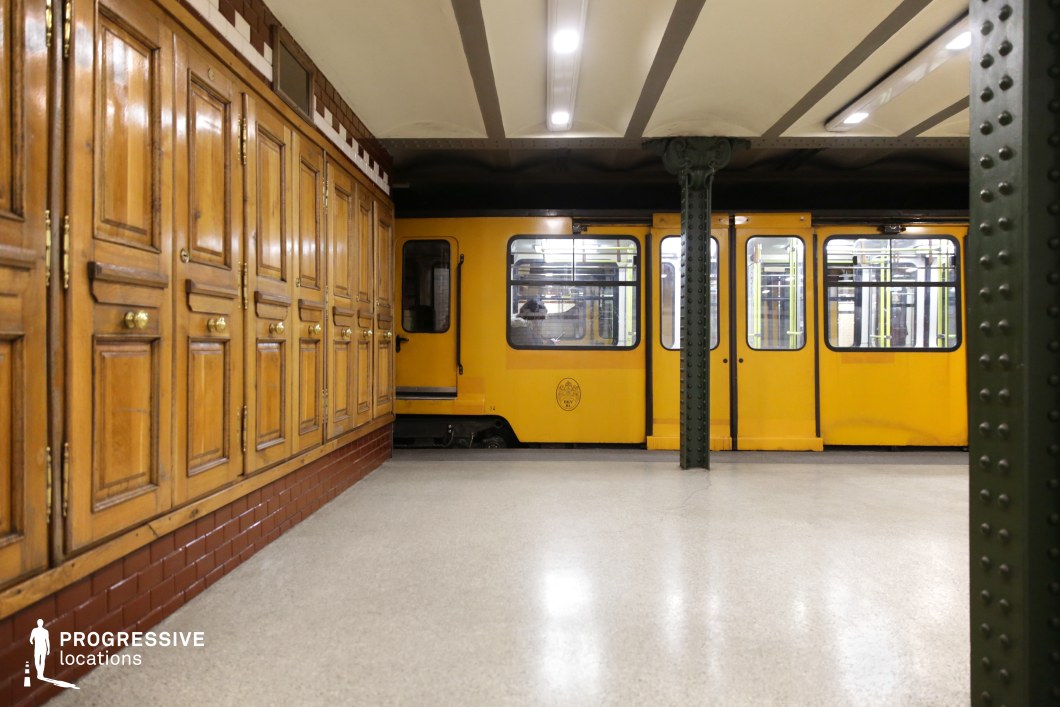 This image showcases a historic underground metro station featuring a striking contrast between a yellow train and a richly detailed wooden-paneled wall. The deep brown cabinetry with brass handles adds a sense of old-world craftsmanship, complemented by red tile accents at its base. Dark green riveted iron columns frame the scene, supporting an exposed-beam ceiling that enhances the station’s vintage character. The polished neutral flooring and soft lighting create a warm yet structured ambiance. This location is ideal for filming period dramas, introspective urban sequences, or atmospheric noir-style storytelling, offering a visually rich transit setting with a nostalgic European feel.