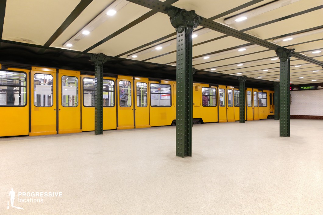 This image depicts a historic underground metro station featuring a striking yellow train alongside a platform supported by dark green riveted iron columns. The station’s ceiling is structured with exposed metal beams, contributing to its industrial-era aesthetic. The flooring is clean and neutral-toned, while the walls are adorned with white tiles and a red accent strip, reinforcing the classic subway design. The scene's open composition and strong linear perspective create a visually compelling setting. This location is ideal for filming period pieces, retro-inspired urban sequences, or suspenseful thriller scenes, offering a timeless underground transit atmosphere reminiscent of early 20th-century European metro systems.