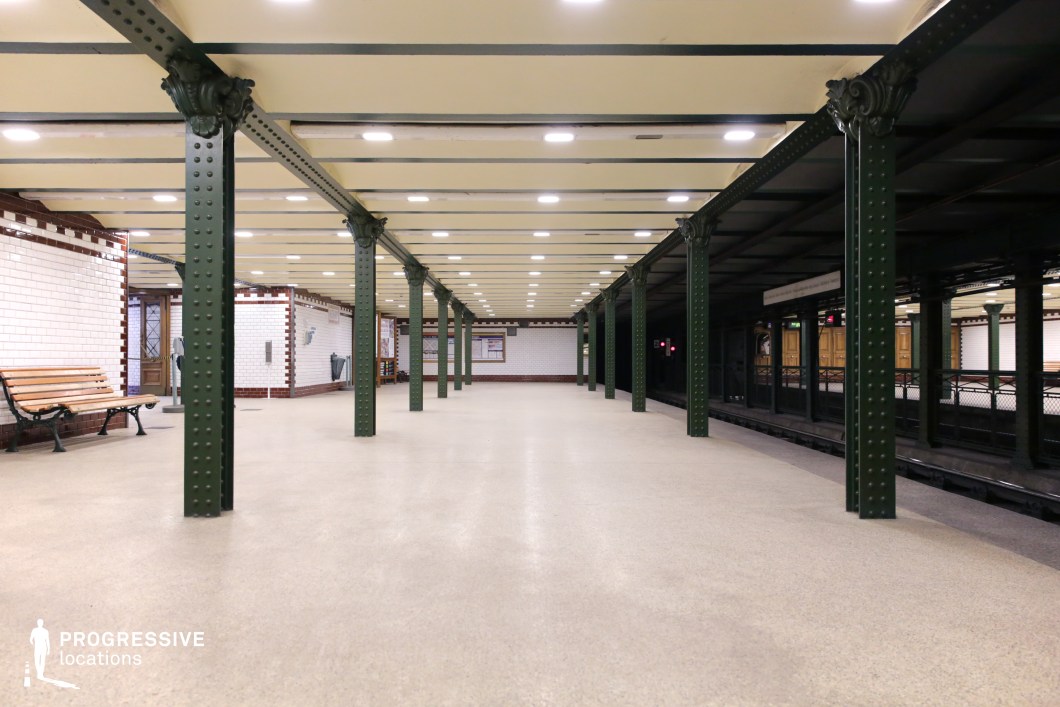 This image showcases a historic underground metro platform with a symmetrical design defined by dark green riveted iron columns supporting an exposed-beam ceiling. The polished neutral flooring reflects the overhead lighting, creating an evenly illuminated space. The walls are adorned with white subway tiles accented by a red trim, while wooden benches and decorative ironwork enhance the classic aesthetic. The depth of the scene, framed by repeating columns, emphasizes the station’s structured elegance. This location is ideal for filming period dramas, noir thrillers, or suspenseful sequences, offering a timeless and atmospheric underground transit setting.