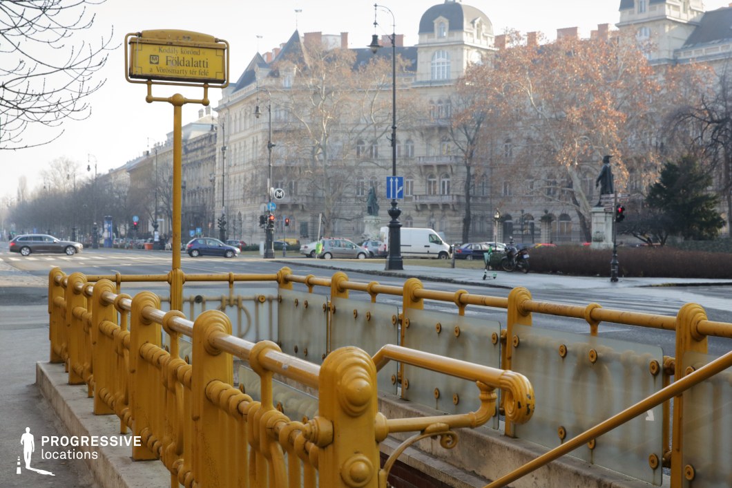 This image captures the entrance to a historic underground metro station, featuring an ornate yellow wrought-iron railing with intricate detailing and frosted glass panels. The entrance descends from a grand boulevard lined with elegant, historic buildings showcasing classic European architecture, complete with decorative facades and mansard roofs. Black cast-iron street lamps and a prominent statue contribute to the refined urban atmosphere. The perspective emphasizes the structured depth of the scene, making this location ideal for filming period dramas, European cityscape sequences, or atmospheric urban scenes with a historic and sophisticated ambiance.