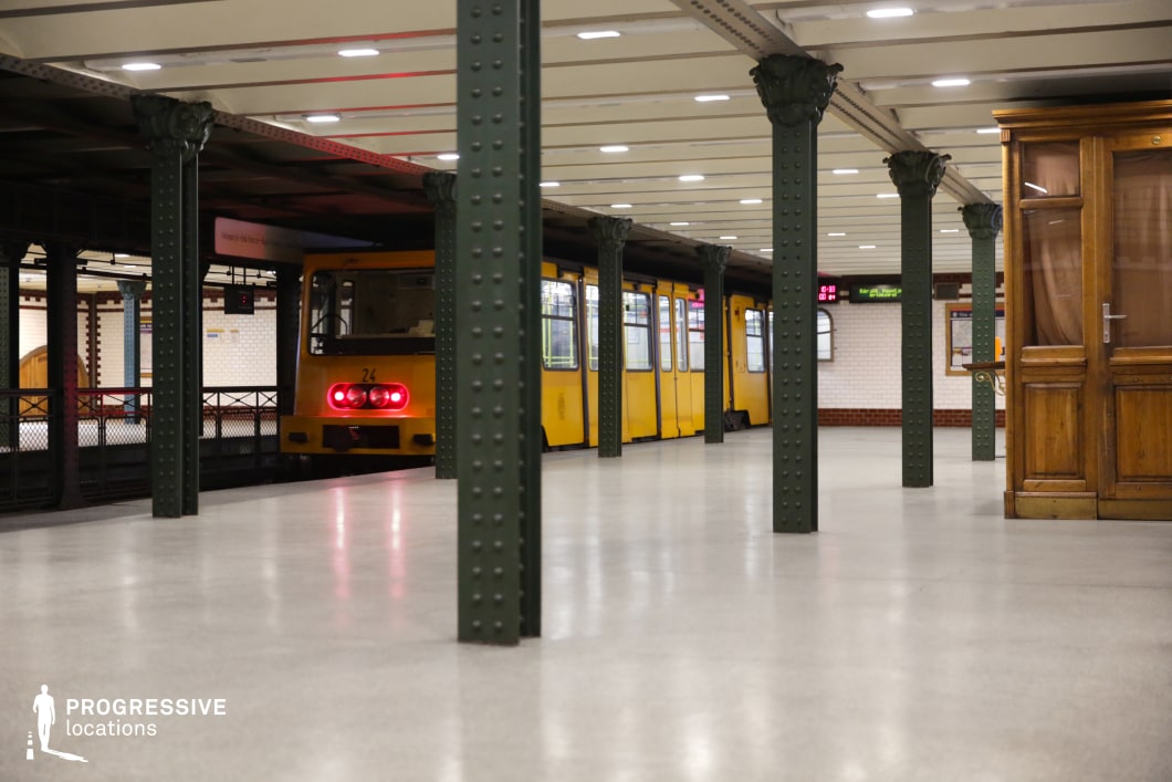 This image captures a historic underground metro platform with a yellow train departing in the background. The setting is defined by dark green riveted iron columns supporting an exposed-beam ceiling, creating a strong industrial-era aesthetic. The polished neutral flooring reflects the station’s overhead lighting, while the wooden ticket booth and paneling add warmth and a classic European charm. White subway tiles with red trim further emphasize the station’s vintage appeal. The combination of structural depth and a receding train makes this an ideal filming location for suspenseful departures, noir-inspired urban sequences, or period dramas set in a timeless transit environment.